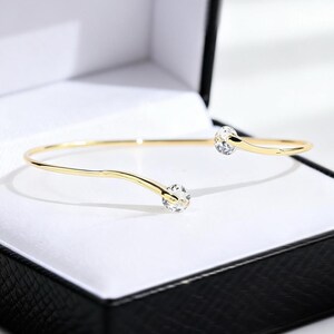 May include: A gold-toned open cuff bracelet featuring a thin, polished metal band that curves slightly at each end. Each terminal of the bracelet is capped with a single, clear, round faceted gemstone that catches the light.