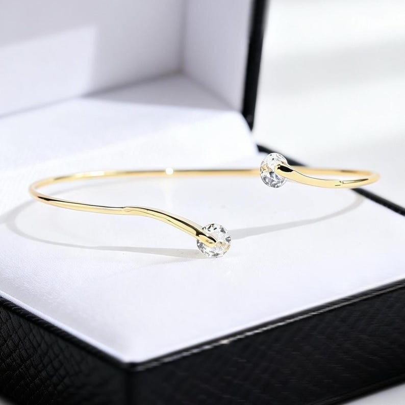 May include: A gold-toned open cuff bracelet featuring a thin, polished metal band that curves slightly at each end. Each terminal of the bracelet is capped with a single, clear, round faceted gemstone that catches the light.