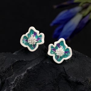 May include: A pair of flower-shaped stud earrings. The earrings have a silver-coloured border, with a teal and purple enamel design. The centre of each earring is adorned with small, clear crystals. The earrings are displayed on a black, textured surface.