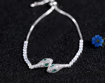 May include: A silver-toned bracelet featuring a snake design. The snake's head and tail are adorned with small, clear stones and green accents. The bracelet is set against a black background, with a small blue flower in the background.