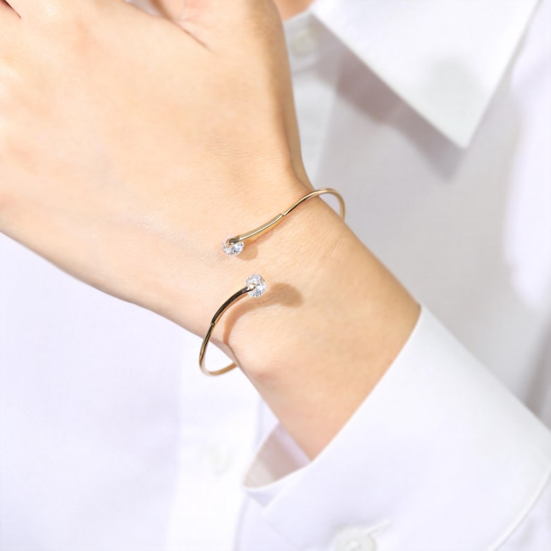 May include: A gold-toned open cuff bracelet featuring two clear, round-cut stones at each terminal end. The thin, polished metal band curves around the wrist, showcasing a minimalist design with sparkling accents at the open gap of the jewellery piece.