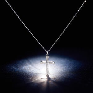 May include: A silver cross pendant necklace. The cross is detailed with small embellishments and a central gemstone. The necklace hangs from a delicate chain, set against a dark background with a spotlight effect.