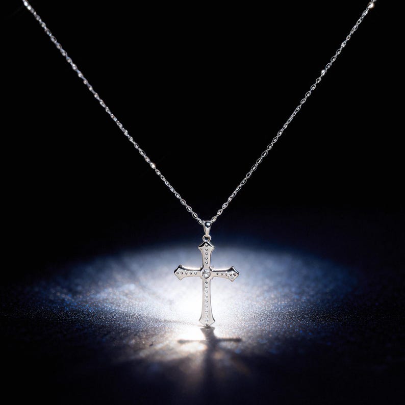May include: A silver cross pendant necklace. The cross is detailed with small embellishments and a central gemstone. The necklace hangs from a delicate chain, set against a dark background with a spotlight effect.