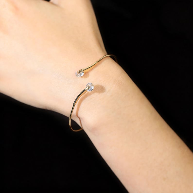 May include: A gold-toned open cuff bracelet featuring a thin, polished metal band. The two ends of the bracelet are finished with small, clear, round faceted stones that provide a subtle sparkle at the opening of the circular wrist accessory.