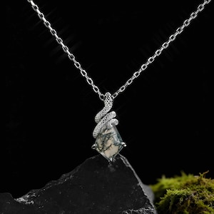 May include: A silver necklace featuring a pendant with a geometric, moss agate stone. A detailed silver snake wraps around the stone. The chain is delicate and the background is black.