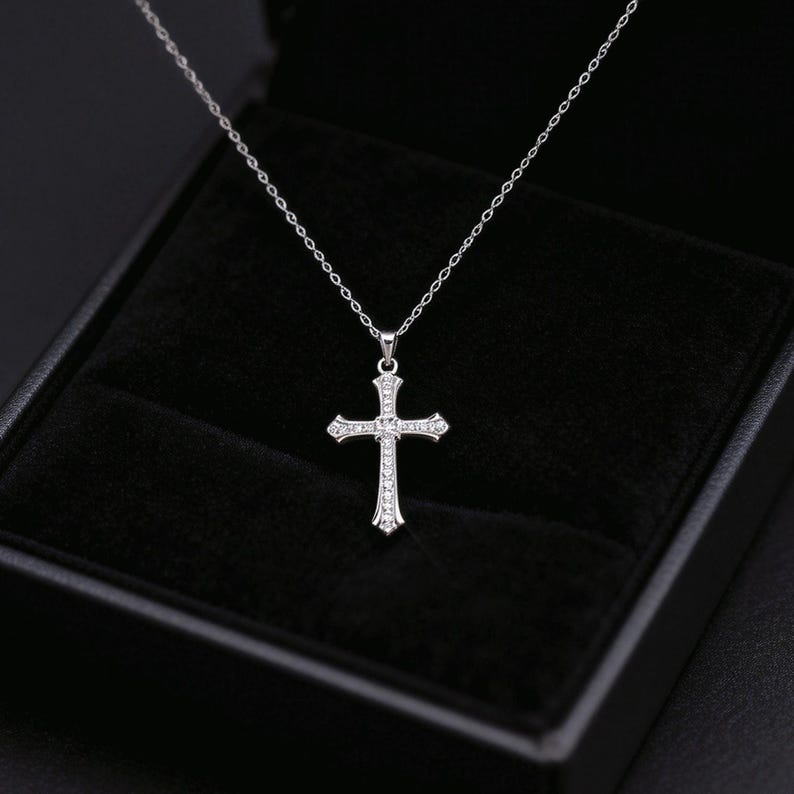 May include: A silver-colored cross pendant necklace with a delicate chain. The cross is adorned with small, sparkling gemstones. The necklace is presented in a black jewelry box.