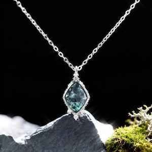 Puede incluir: Collar de plata con un colgante en forma de diamante. El colgante presenta una piedra preciosa de color verde azulado con un marco plateado decorativo y pequeños detalles blancos. La cadena del collar también es plateada.