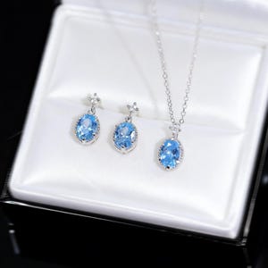 May include: A jewelry set featuring a necklace and matching earrings. The set includes a silver chain necklace with a pendant and a pair of earrings, each with a light blue oval gemstone. The jewelry is displayed in a white box.