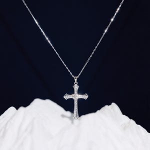 May include: A silver-colored necklace with a cross pendant. The cross is adorned with small, clear gemstones and a larger gemstone at the center. The necklace hangs against a dark background and rests on a white, textured surface.