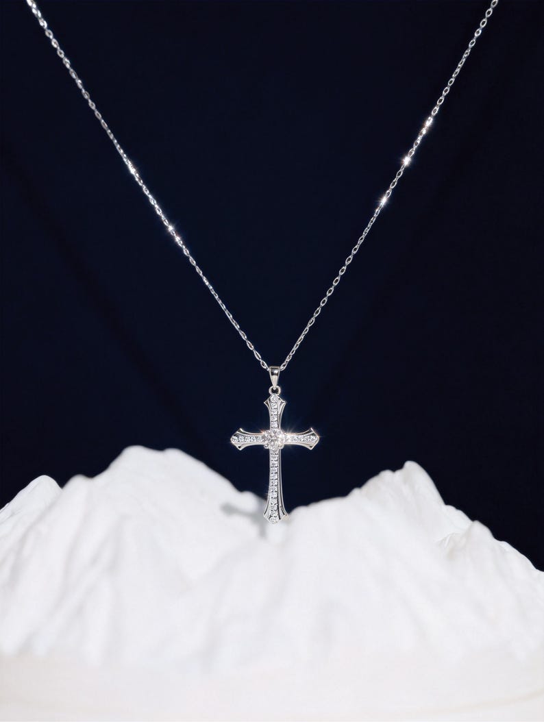 May include: A silver-colored necklace with a cross pendant. The cross is adorned with small, clear gemstones and a larger gemstone at the center. The necklace hangs against a dark background and rests on a white, textured surface.
