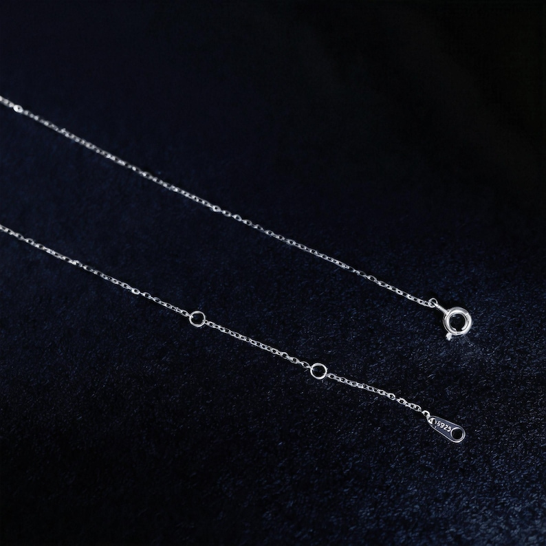 May include: A delicate silver necklace with a fine chain. The necklace features a small clasp and adjustable length with several small rings. The chain is displayed against a dark background.