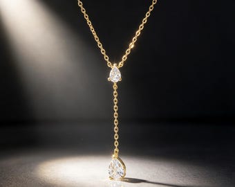 May include: A gold-toned Y-shaped necklace featuring a delicate chain with two clear, pear-shaped faceted stones. One stone is positioned at the center junction, and the second stone hangs at the end of the vertical drop chain segment.