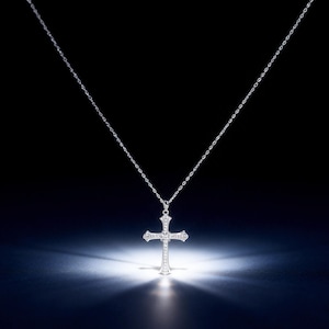 May include: A silver cross pendant necklace. The cross is encrusted with small, sparkling stones and hangs from a delicate chain. The necklace is displayed against a dark background with a spotlight illuminating the cross.