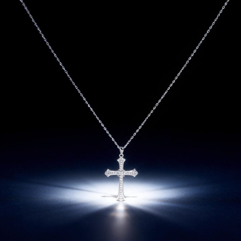May include: A silver cross pendant necklace. The cross is encrusted with small, sparkling stones and hangs from a delicate chain. The necklace is displayed against a dark background with a spotlight illuminating the cross.