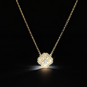 Peut inclure: Un collier doré avec un pendentif en forme de trèfle à quatre feuilles. Le pendentif présente un centre blanc avec un diamant, entouré de petits diamants. La chaîne est délicate et se détache sur un fond noir.
