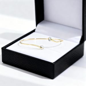 May include: A gold open cuff bracelet featuring two clear, round, faceted stones at each end. The bracelet is presented inside a black jewellery box with a white interior lining, showcasing a simple and elegant design with a polished metal finish.