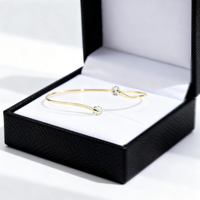 May include: A gold open cuff bracelet featuring two clear, round, faceted stones at each end. The bracelet is presented inside a black jewellery box with a white interior lining, showcasing a simple and elegant design with a polished metal finish.