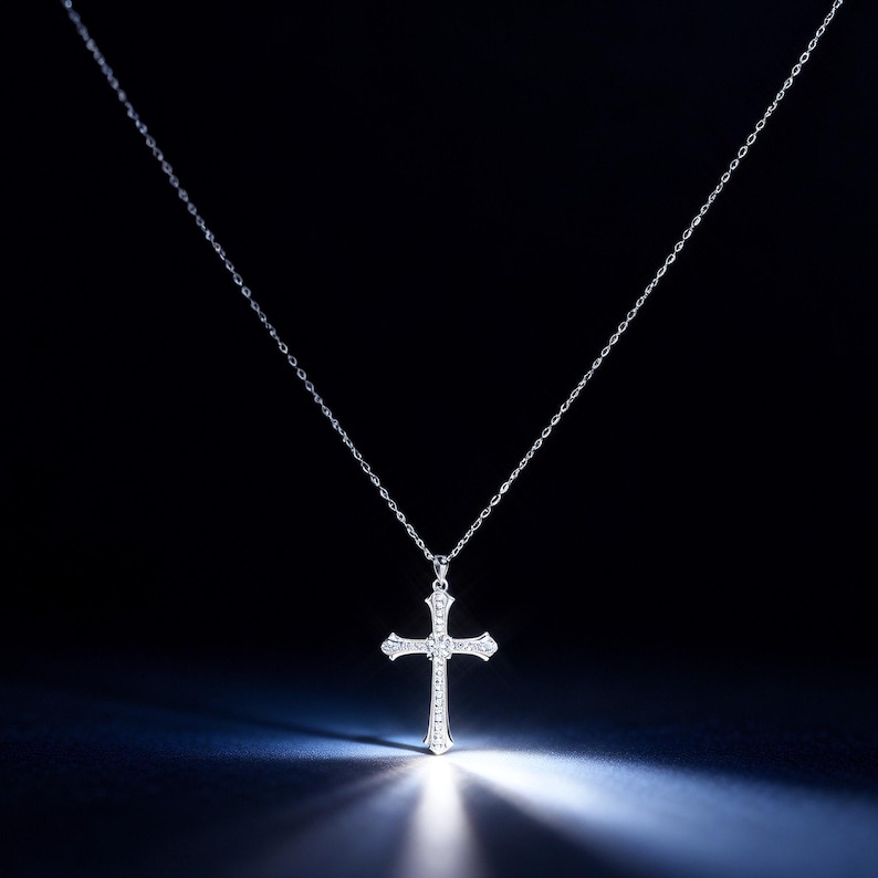 May include: A silver cross pendant necklace with a delicate chain. The cross is adorned with small, sparkling gemstones. The necklace is displayed against a dark background, with a spotlight illuminating the cross.