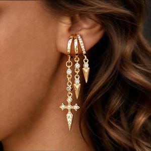 May include: Three gold-toned hoop earrings with clear stones. The first features a long chain with a cross pendant. The second has a dangling spike charm. The third is a small hoop with a shorter spike charm. All pieces are set with small clear crystals.