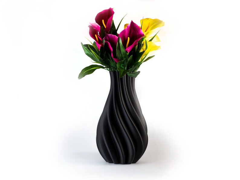 Puede incluir: Un jarr&oacute;n negro texturizado con un dise&ntilde;o en espiral contiene un ramo de flores coloridas. Las flores incluyen calas de color magenta oscuro y amarillo con follaje verde. El jarr&oacute;n est&aacute; sobre un fondo blanco.