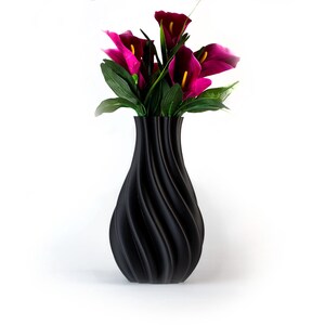 Puede incluir: Un jarr&oacute;n negro decorativo con un dise&ntilde;o en espiral, que contiene un ramo de calas magenta y hojas verdes. El jarr&oacute;n tiene un acabado liso y mate y est&aacute; sobre un fondo blanco.
