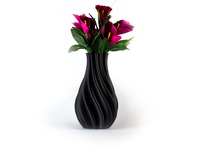 Puede incluir: Un jarr&oacute;n negro decorativo con un dise&ntilde;o en espiral, que contiene un ramo de calas magenta y hojas verdes. El jarr&oacute;n tiene un acabado liso y mate y est&aacute; sobre un fondo blanco.