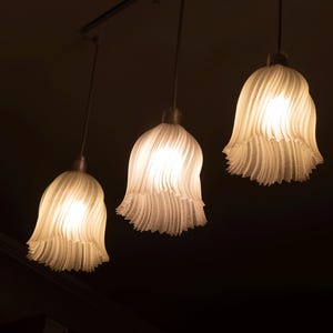 May include: Three pendant lights with white, textured, bell-shaped shades hang from a dark ceiling. The shades have a ruffled, layered design, and each contains a bright, warm light. The overall effect is soft and inviting.