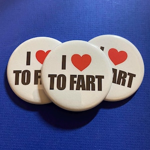 Puede incluir: Tres botones redondos blancos con el texto "I ❤️ TO FART" en negro y un corazón rojo. Los botones están colocados sobre una superficie azul.