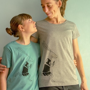 Pode incluir: Duas pessoas vestindo camisetas com um gráfico de um coelho correndo. O coelho está vestindo uma camiseta sem mangas e shorts. A pessoa à esquerda está vestindo uma camiseta turquesa e a pessoa à direita está vestindo uma camiseta cinza.