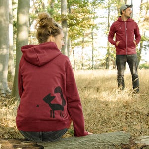 Könnte beinhalten: Burgunderfarbener Kapuzenpullover mit einem schwarzen Fuchsgrafik auf dem Rücken. Der Pullover hat einen durchgehenden Reißverschluss und eine Kapuze. Eine Person trägt den Pullover in einer Waldumgebung.