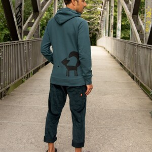 May include: A teal blue hooded sweatshirt with a black fox graphic on the back. The sweatshirt is worn by a person standing on a bridge.