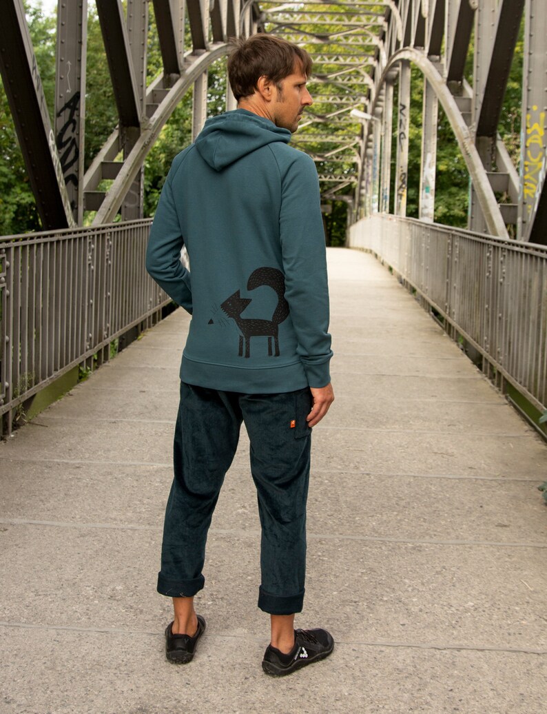 May include: A teal blue hooded sweatshirt with a black fox graphic on the back. The sweatshirt is worn by a person standing on a bridge.