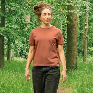 May include: A rust-colored short-sleeve t-shirt. The shirt has a crew neck and a simple, classic design. The person is wearing black jeans and is walking on a path in a forest.