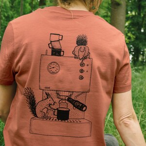 May include: Rust-colored t-shirt featuring a black and white illustration of a coffee machine with a squirrel and a hedgehog. The squirrel is holding a cup, and the hedgehog is perched on top. A stack of coffee cups is also visible.