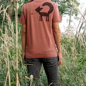 May include: A woman wearing a rust-coloured t-shirt with a black silhouette of a fox on the back. She is standing in a field of tall grass.