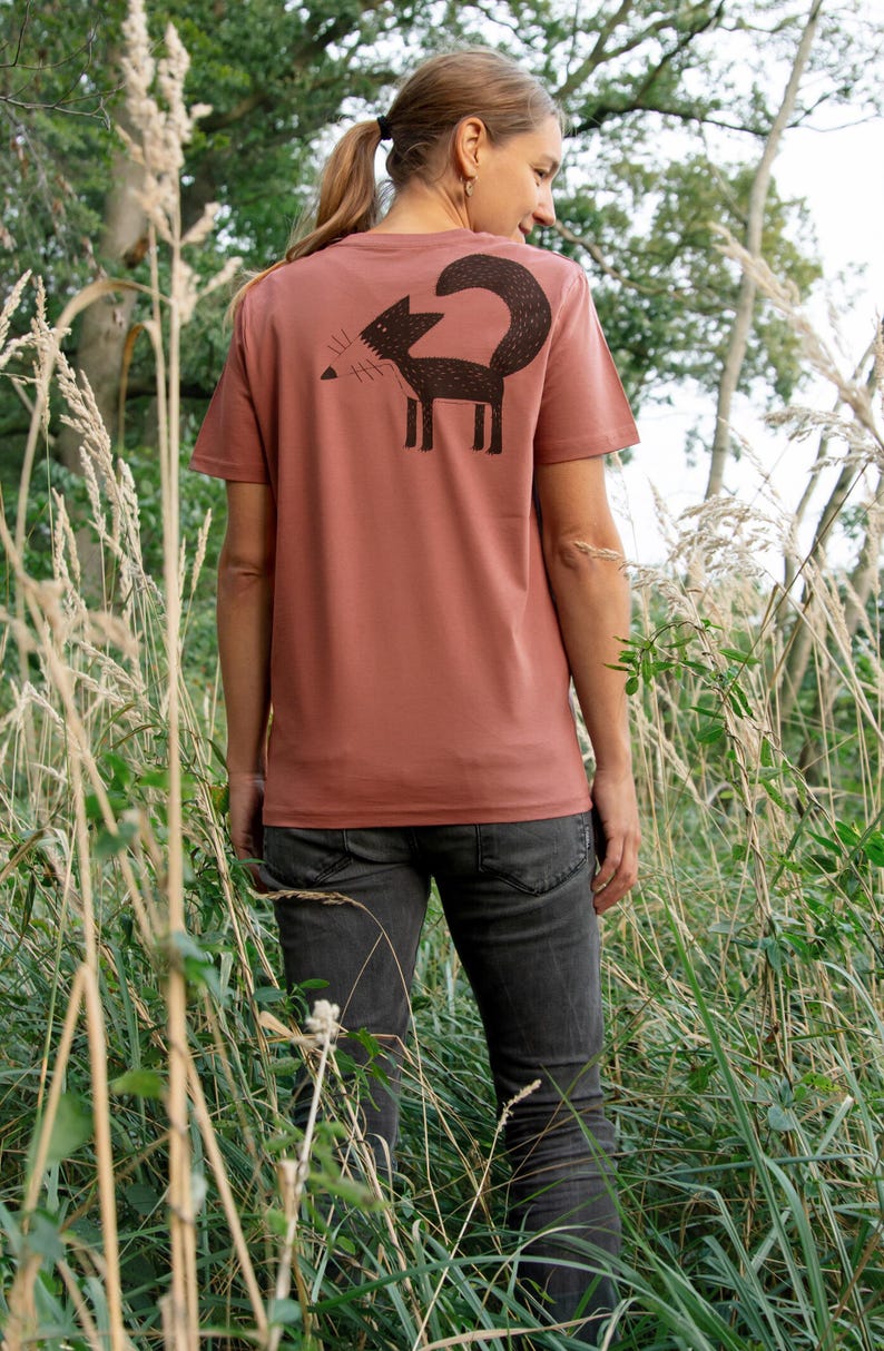 May include: A woman wearing a rust-coloured t-shirt with a black silhouette of a fox on the back. She is standing in a field of tall grass.