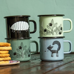 May include: Four enamel mugs with black and white illustrations. The mugs are stacked in two pairs. The top mug in the left stack features a black and white illustration of a pill bug. The top mug in the right stack features a black and white illustration of a plant with berries. The bottom mug in the left stack features a black and white illustration of a plant with berries. The bottom mug in the right stack features a black and white illustration of a fox.
