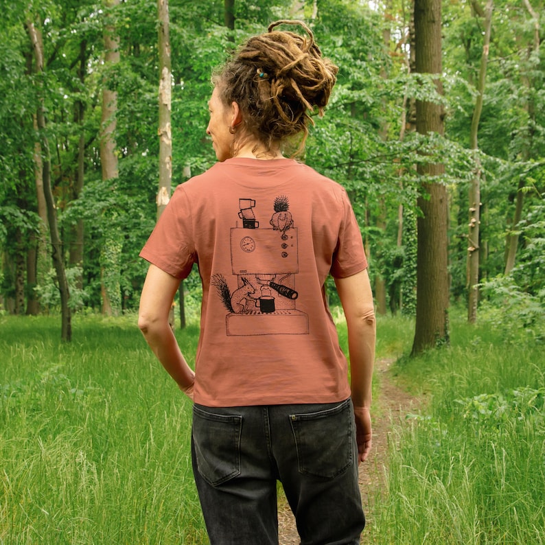 May include: A rust-colored t-shirt with a black graphic of a coffee machine, a squirrel, and a small figure. The t-shirt is short-sleeved and is being worn outdoors in a forest setting. The person is wearing black jeans.