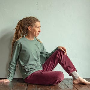 May include: A person wearing a sage green long-sleeved shirt with a black floral print and maroon trousers. The person is sitting on a wooden floor with their legs crossed.