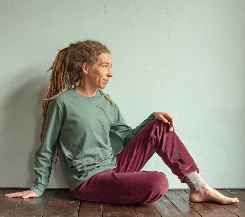 May include: A person wearing a sage green long-sleeved shirt with a black floral print and maroon trousers. The person is sitting on a wooden floor with their legs crossed.