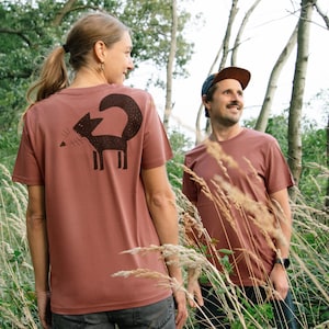 May include: Two people wearing brown t-shirts with a black fox graphic on the back. The person on the left is a woman with long brown hair and the person on the right is a man wearing a brown cap. They are standing in a field of tall grass.