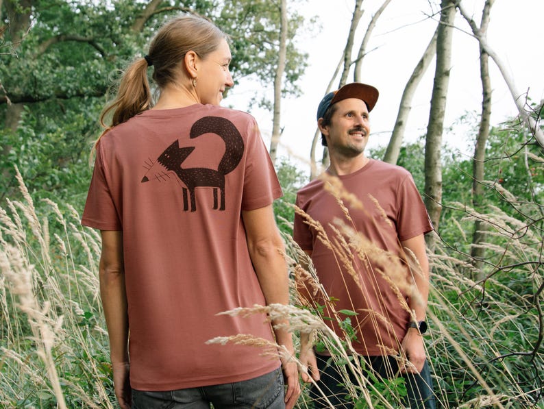 May include: Two people wearing brown t-shirts with a black fox graphic on the back. The person on the left is a woman with long brown hair and the person on the right is a man wearing a brown cap. They are standing in a field of tall grass.