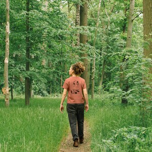 May include: A person wearing a rust-colored t-shirt with a black graphic design, walking on a path through a lush green forest. The t-shirt features a minimalist illustration. The person is wearing dark pants and brown shoes.