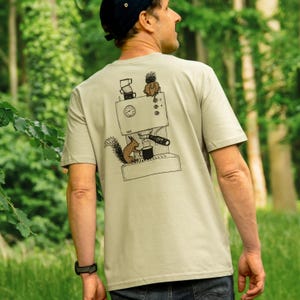 Könnte beinhalten: Beiges T-Shirt mit einer Schwarz-Weiß-Illustration einer Kaffeemaschine und einem Eichhörnchen. Das Eichhörnchen hält eine Kaffeetasse. Das T-Shirt ist kurzärmlig und hat einen Rundhalsausschnitt.