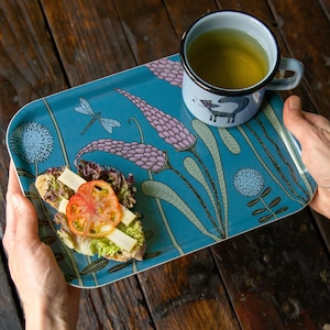 Pode incluir: Uma bandeja de servir turquesa com um design floral, contendo uma sanduíche de tomate e queijo e uma caneca de chá. A caneca tem uma borda preta e uma ilustração de raposa. O design da bandeja inclui libélulas e flores estilizadas.