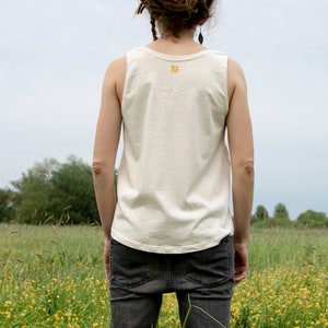 Puede incluir: Un top blanco sin mangas con un peque&ntilde;o logotipo en la espalda. La persona que lleva el top tiene rastas y est&aacute; de pie en un campo de flores amarillas.