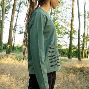 May include: A woman wearing a long-sleeved, teal green shirt with a black floral print. The shirt has a relaxed fit and a crew neckline. The woman is standing in a woodland setting.