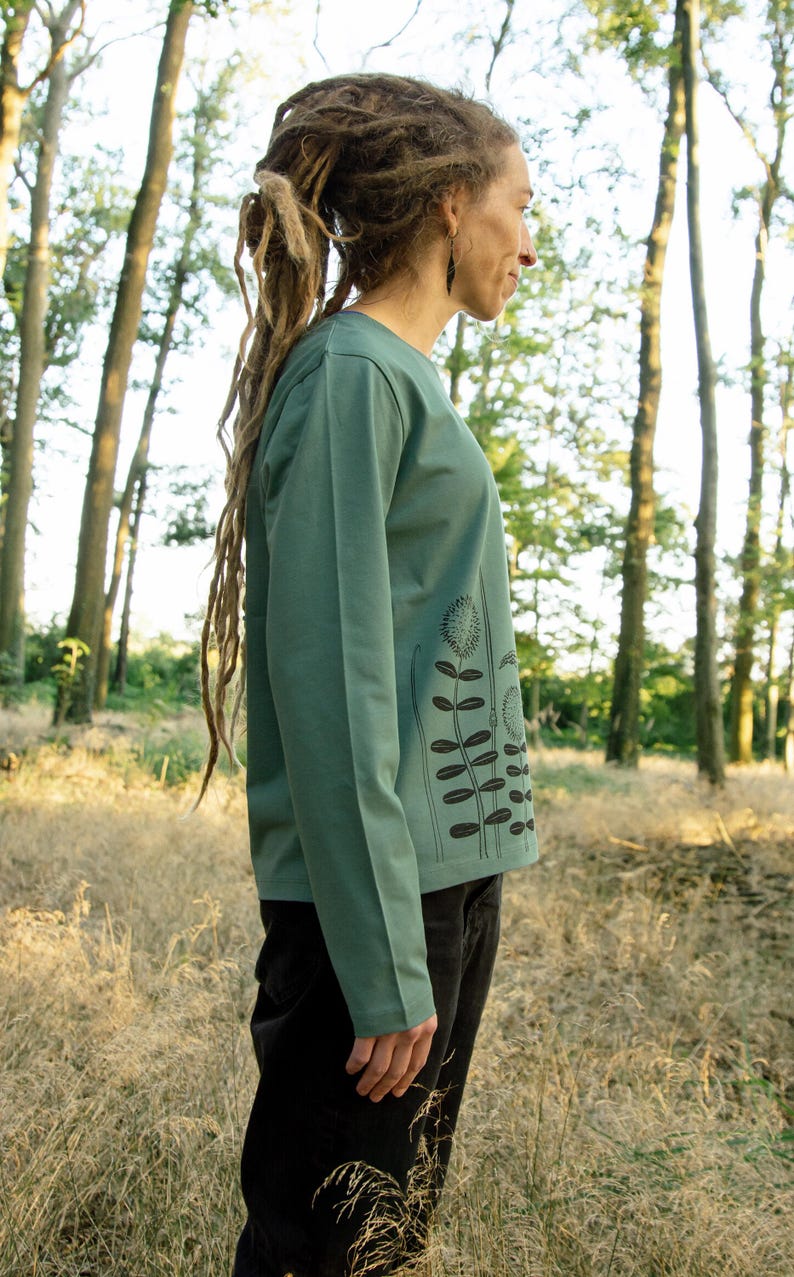 May include: A woman wearing a long-sleeved, teal green shirt with a black floral print. The shirt has a relaxed fit and a crew neckline. The woman is standing in a woodland setting.
