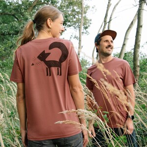 May include: Two people wearing brown t-shirts with a black fox graphic on the back. The fox is stylised and has a long tail. The people are standing in a field of tall grass.