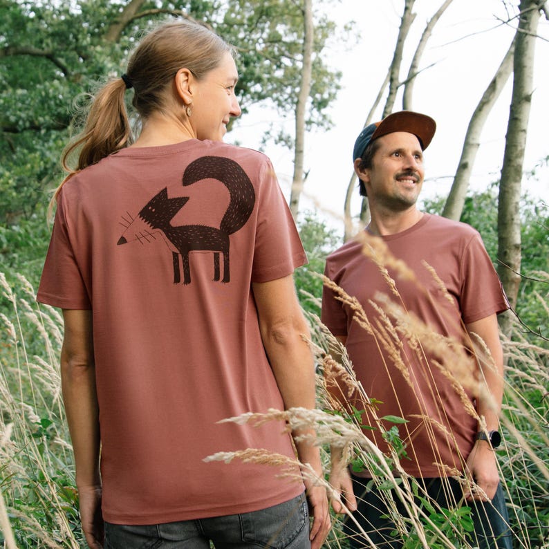 May include: Two people wearing brown t-shirts with a black fox graphic on the back. The fox is stylised and has a long tail. The people are standing in a field of tall grass.